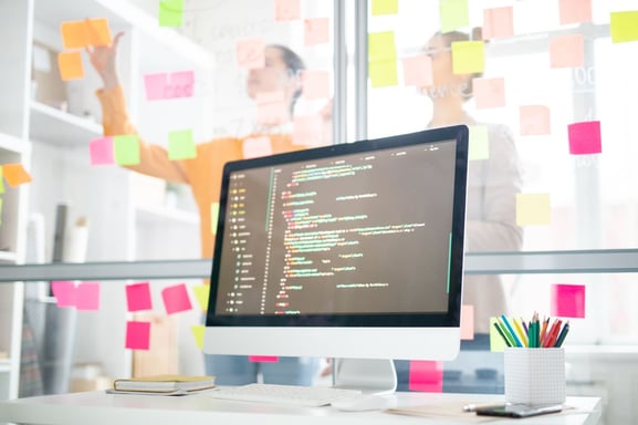 A computer screen with code sitting on a desk in front of a window with brainstorming sticky notes, demonstrating ease of use in software implementation